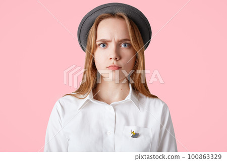 Candid shot of grumpy sad serious female wears elegant black hat and white blouse, looks directly into camera, isolated over pink background. Elegant grumpy woman. Facial expressions concept Candid shot of grumpy sad serious female wears elegant black hat and white blouse, looks directly into camera, isolated over pink background. Elegant grumpy woman. Facial expressions concept 100863329