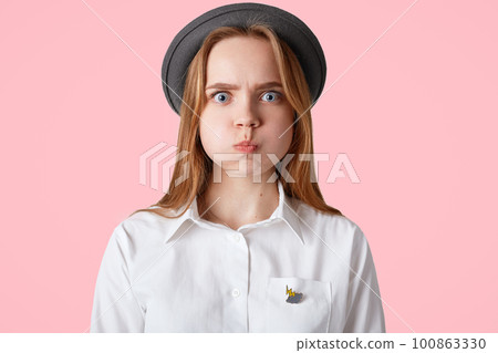 Shot of annoyed blue eyed female with pleasant appearance, blows cheeks, wears elegant black hat and white shirt, has grumpy expression, poses against pink background. Facial expressions concept Shot of annoyed blue eyed female with pleasant appearance, blows cheeks, wears elegant black hat and white shirt, has grumpy expression, poses against pink background. Facial expressions concept 100863330