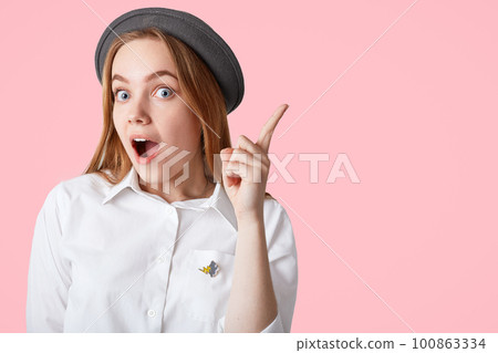 Surprised beautiful European female keeps fore finger raised, wears elegant shirt and hat, gets good idea to put in life, isolated over pink background with copy space for your promotional content Surprised beautiful European female keeps fore finger raised, wears elegant shirt and hat, gets good idea to put in life, isolated over pink background with copy space for your promotional content 100863334