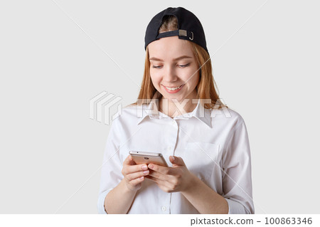 Close up portrait of smiling beautiful teenager with visor cap back, has brown hair, poses at white studio wall, holds smartphone, checking social network, comunicates with friens, wears white blouse. Close up portrait of smiling beautiful teenager with visor cap back, has brown hair, poses at white studio wall, holds smartphone, checking social network, comunicates with friens, wears white blouse. 100863346