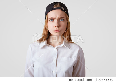Waist up portrait of grumpy female model with sullen expression, wears black cap and elegant shirt, being dissatisfied with something, isolated over white studio background. People and emotions Waist up portrait of grumpy female model with sullen expression, wears black cap and elegant shirt, being dissatisfied with something, isolated over white studio background. People and emotions 100863350