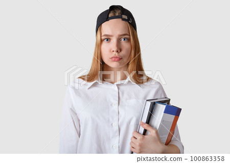 Young student has bad result of exam, posing with serious and upset facial expression. Schoolgirl stands isolated over white background, holding paper folders in hands, wearing blouse. Copy space. 100863358