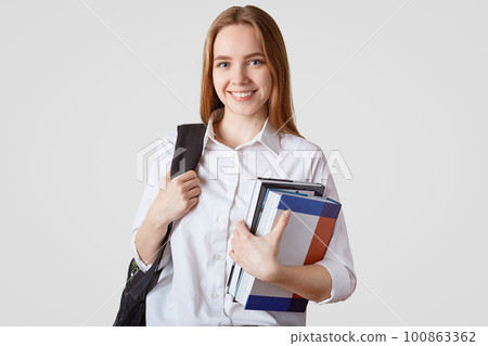 Back to school concept. Smiling European schoolgirl in elegant clothes, carries rucksack and pile of books, glad to have weekend after long tired week of studying, isolated over white background Back to school concept. Smiling European schoolgirl in elegant clothes, carries rucksack and pile of books, glad to have weekend after long tired week of studying, isolated over white background 100863362