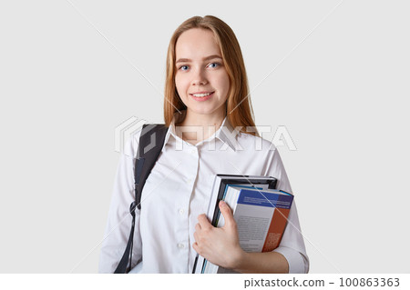Close up portraitof young girl teenagercarries rucksack, holds colourful folder with papers, has pleasant smile, has lectures in university, poses isolated on yellow background with copy space. Close up portraitof young girl teenagercarries rucksack, holds colourful folder with papers, has pleasant smile, has lectures in university, poses isolated on yellow background with copy space. 100863363