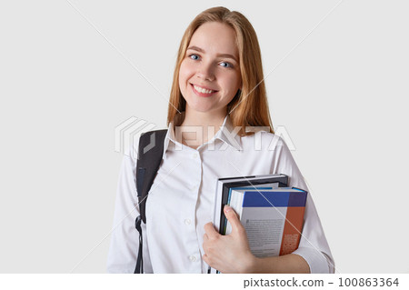 Indoor shot of beautiful teenager girl with black backpack and paper folder in hands, looking happy, attractive student woman isolated on white background. Teenagers, education and success concept. Indoor shot of beautiful teenager girl with black backpack and paper folder in hands, looking happy, attractive student woman isolated on white background. Teenagers, education and success concept. 100863364