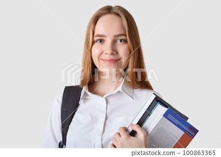 Sweet adorable blond haired young female poses isolated over white background, holding many books in both hands, looking directly at camera, looks pleased and confident, having black bagback. Sweet adorable blond haired young female poses isolated over white background, holding many books in both hands, looking directly at camera, looks pleased and confident, having black bagback. 100863365