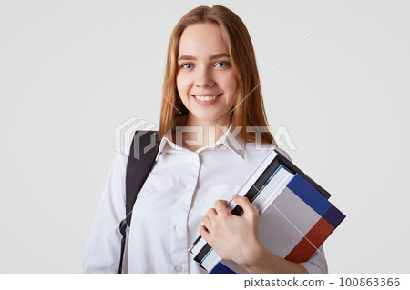 Adorable school girl with blue eyes, shining smile, wears elegant white shirt, carries books and rucksack, has long hair, prepares for exam, isolated over studio wall. Back to school concept Adorable school girl with blue eyes, shining smile, wears elegant white shirt, carries books and rucksack, has long hair, prepares for exam, isolated over studio wall. Back to school concept 100863366