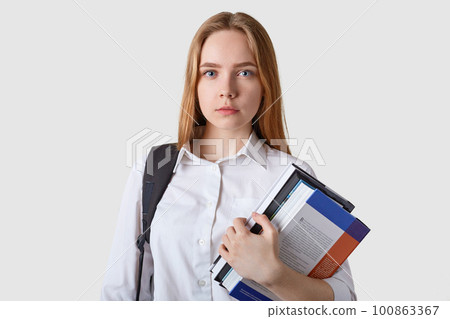 Studio of serious caucasian fair haired young woman with paper folder in hands, wearing white blouse, looking at camera with strict expression, model posing against studio background. Student concept. 100863367