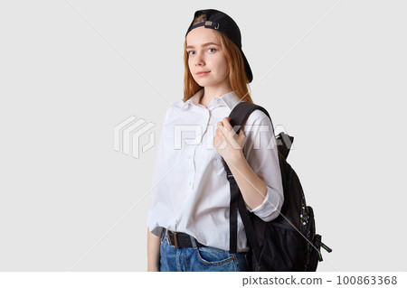 Slender beautiful energetic school girl goes to school wearing blue jeans, white shirt, black cap and bagpack, looks concentrated on studying, holidays come to end. Studying and education concept. Slender beautiful energetic school girl goes to school wearing blue jeans, white shirt, black cap and bagpack, looks concentrated on studying, holidays come to end. Studying and education concept. 100863368