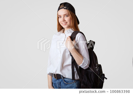 Happy schoolgirl in black cap and white shirt, carries rucksack with books, ready for starting new studying year, poses against white background. College student returns back home from classes Happy schoolgirl in black cap and white shirt, carries rucksack with books, ready for starting new studying year, poses against white background. College student returns back home from classes 100863398
