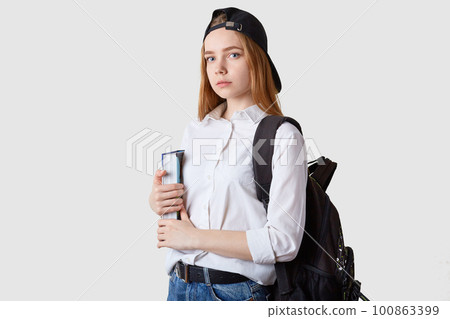 Horizontal shot of student girl standing isolated over white background, holding paper folders in hands, wearing blouse, black cap and backpack, looking at camera. Education and teenager concept. Horizontal shot of student girl standing isolated over white background, holding paper folders in hands, wearing blouse, black cap and backpack, looking at camera. Education and teenager concept. 100863399