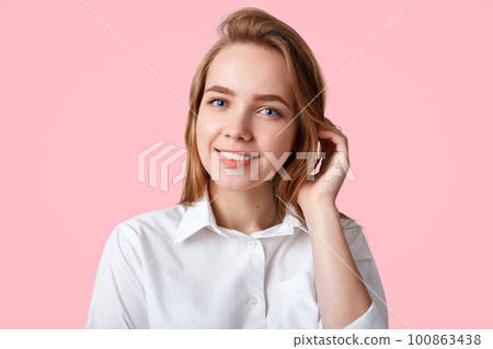 Headshot of adorable female college student with blue eyes, healthy pure skin, wears white shirt, poses against pink background, has gentle pleasant smile. People, positive emotions concept. Headshot of adorable female college student with blue eyes, healthy pure skin, wears white shirt, poses against pink background, has gentle pleasant smile. People, positive emotions concept. 100863438