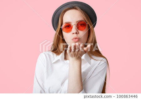 Lovely fashionable hipster girl wears trendy pink shades and black hat, blows air kiss at camera, poses against pink background, expresses her love to surrounding, Pleasant looking female flirts Lovely fashionable hipster girl wears trendy pink shades and black hat, blows air kiss at camera, poses against pink background, expresses her love to surrounding, Pleasant looking female flirts 100863446