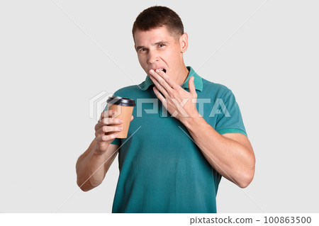 Tired guy yawns with sleepy expression, covers mouth with palm, holds disposable paper cup of coffee, dressed in casual t shirt, poses against white background. Morning time. Coffee break concept Tired guy yawns with sleepy expression, covers mouth with palm, holds disposable paper cup of coffee, dressed in casual t shirt, poses against white background. Morning time. Coffee break concept 100863500