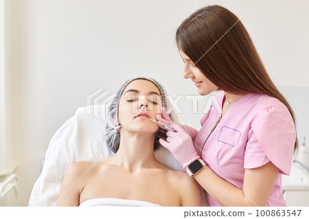Woman receiving anti ageing injection in her face at cosmetology salon while sitting on couh. Beauty woman giving injections. Face aging injection. Aesthetic medicine, cosmetology, skin care concept. 100863547