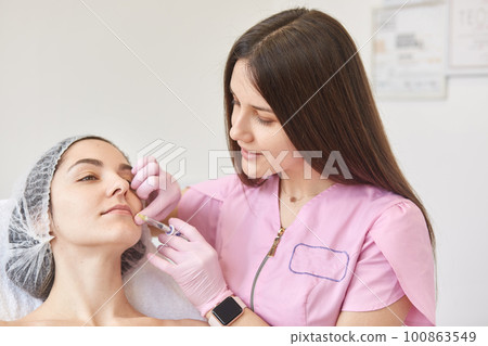 Cosmetologist making injection in female lips with syringe. Young beautiful woman is receiving injection in cosmetology salon, looks calm, wants to have plump lips. Aesthetic medicine concept. 100863549
