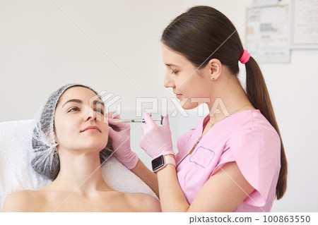 Patient attractive client lies in cosmetic chair, being calm, professional beautician with smart watch wearing pink lab coat, holding syringe, making facial beauty injections. Cosmetology concept. Patient attractive client lies in cosmetic chair, being calm, professional beautician with smart watch wearing pink lab coat, holding syringe, making facial beauty injections. Cosmetology concept. 100863550