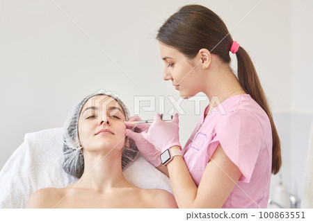 Photo of young oman getting collage injection in her face at clinic. Cosmetologist wears pink medical gown filling female wrinkles by hyaluronic acid. Woman sits on couh calmly. Anti aging concept. 100863551