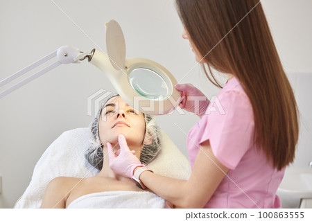 Experienced young cosmetologist in pink lab coat holding magnifying glass in one hand, switching on lamp, touching womans face, assessing results, working in beauty parlor. Cosmetology concept. Experienced young cosmetologist in pink lab coat holding magnifying glass in one hand, switching on lamp, touching womans face, assessing results, working in beauty parlor. Cosmetology concept. 100863555