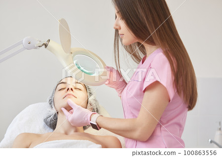 Cosmetologist dresses pink madical gown, latex gloves examines patient face, finds defects under lamp, prepering woman for ejuvenation and hydration procedures. Cosmetology, aethetic medicine concept. Cosmetologist dresses pink madical gown, latex gloves examines patient face, finds defects under lamp, prepering woman for ejuvenation and hydration procedures. Cosmetology, aethetic medicine concept. 100863556