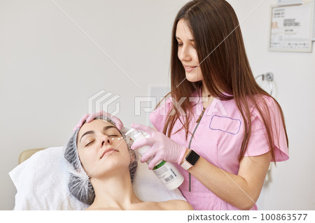 Smiling pleasant beautician in pink lab coat and watch on wrist standing near her client, touching her forehead, holding bottle with moisturizing cream, applying on face, having positive expression. Smiling pleasant beautician in pink lab coat and watch on wrist standing near her client, touching her forehead, holding bottle with moisturizing cream, applying on face, having positive expression. 100863577