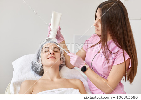 Doctor cosmetologist doing face lifting massage for young brunette woman, client lying on couh woth closed eyes, wearing white towel and medical cap. Aethsetic medicine, cosmetology and beauty concept 100863586