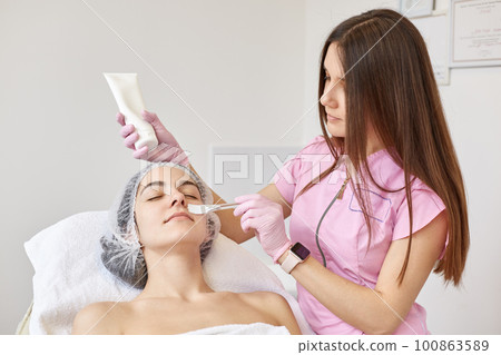 Peaceful client with closed eyes had beauty procedures, laying calm on cosmetic chair, having medical cap on head. Beautician working in beauty parlor, holding tube of cream, applying it with brush. 100863589