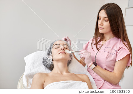 Beautiful young lady having face lifting massage, brunette woman lying on couch with closed eyes, wearing white towel and medical cap. Doctor cosmetologist working. Cosmetology and beauty concept 100863590