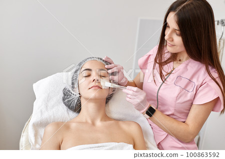 Horizontal shot of oman being at cosmetology salon, sitting on couch, doctor cosmetologist making cleansing procedures, applaying cream on face. Aesthetic medicine, cosmetology, skin care concept. 100863592