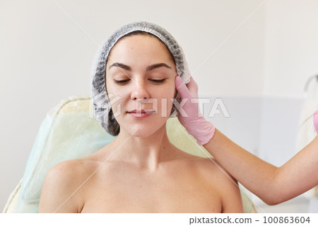 Image of faceless cosmetologist in pink medical gloves at work in cosmetology salon examines attractive woman sits on couh in disposable cap , looking down. Cosmetology and aethetic medicine concept. Image of faceless cosmetologist in pink medical gloves at work in cosmetology salon examines attractive woman sits on couh in disposable cap , looking down. Cosmetology and aethetic medicine concept. 100863604
