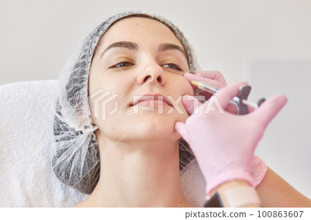 Hands of cosmetologist making injection in face. Young woman gets injections in salon. Face aging, rejuvenation and hydration procedures. Skin care, anti aging and aethetic cosmetology concept. 100863607