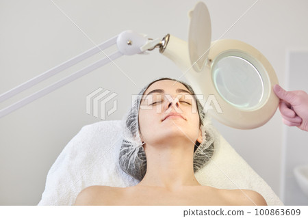 Portrait of delighted client in beauty salon laying in cosmetic chair, relaxing after non surgical beauty procedures, having her eyes closed. Professional looks at skin condition through equipment. 100863609