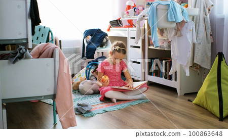 Little girl in pink dress reading book and eating apple in messy room at home Little girl in pink dress reading book and eating apple in messy room at home 100863648