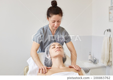 Cosmetologist looks at her client, putting her hands on shoulders, making beauty procedures. Calm model laying on cosmetic chair with closed eyes, waiting for facial massage. Skin care concept. 100863762