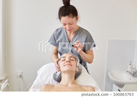 Positive beautician works at beauty parlor. having client, applying moisturizing face oil, preparing face for further procedures. Relaxed smiling woman having rest at spa salon. Skin and care concept. Positive beautician works at beauty parlor. having client, applying moisturizing face oil, preparing face for further procedures. Relaxed smiling woman having rest at spa salon. Skin and care concept. 100863763