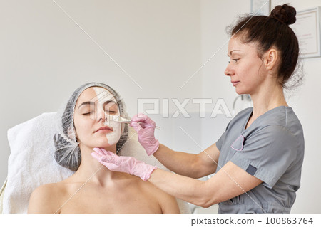 Cosmetologist makes procedure of cleansing and moisturizing of skin, applying mask with special brush to face of woman. Young girl takes care of her skin, visits cosmetology clinic. Aethetic medicine Cosmetologist makes procedure of cleansing and moisturizing of skin, applying mask with special brush to face of woman. Young girl takes care of her skin, visits cosmetology clinic. Aethetic medicine 100863764