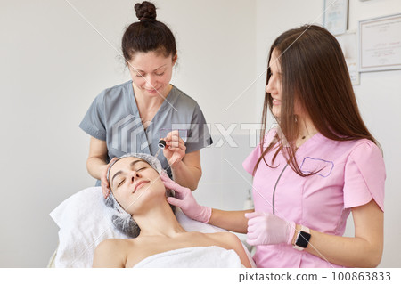 Indoor shot of beautician and her young assistant providing quality customer service to their client laid with closed eyes, preparing skin for cosmetological procedures, applying hydrating oil. 100863833