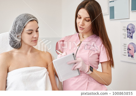 Woman wearing dosposable medical cap being at consultation in cosmetology clinic. Attractive female doctor talking with patient and showing notes. Planning beauty treatment. Aethetic medicine concept. Woman wearing dosposable medical cap being at consultation in cosmetology clinic. Attractive female doctor talking with patient and showing notes. Planning beauty treatment. Aethetic medicine concept. 100863855