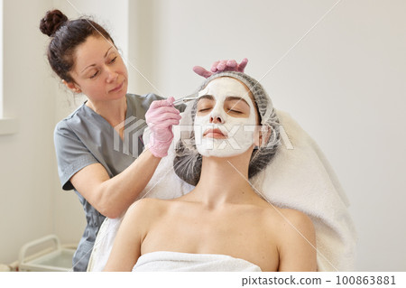 Spa beauty treatment, skincare concept. Woman getting facial care by beautician at spa salon, cosmetologist applays face peeling mask with special brush, female relaxes while sitting on couh. Spa beauty treatment, skincare concept. Woman getting facial care by beautician at spa salon, cosmetologist applays face peeling mask with special brush, female relaxes while sitting on couh. 100863881