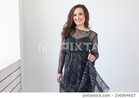 Attractive woman with long brunette hair dressed in fashionable black dress smiling and posing. Laughing female model standing against white wall on background. Attractive woman with long brunette hair dressed in fashionable black dress smiling and posing. Laughing female model standing against white wall on background. 100864687
