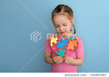 A cute child looks at a postcard made of paper on the day of awareness about autism. Heart-shaped postcard with multicolored puzzles 100865065
