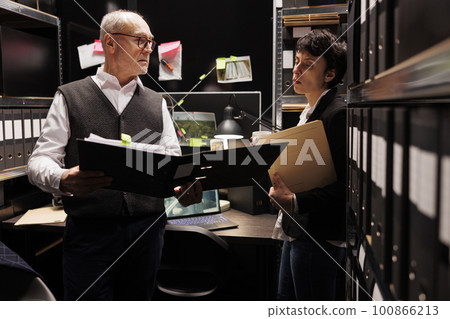 Private detectives discussing missing person evidence, working overtime at mysterious criminal case in arhive room. Police officer checking criminology department report, reading confidential files 100866213