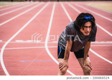 Male runner gasping for breath while slouching on an athletics track 100868524
