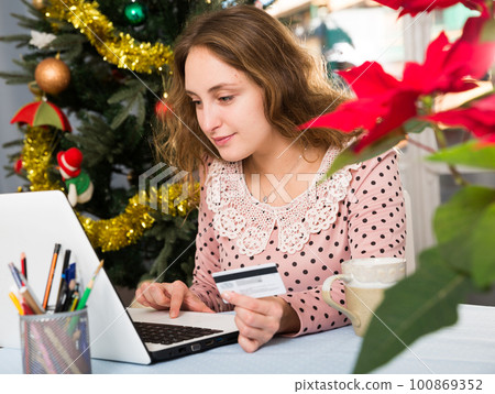 Happy young woman with credit card and laptop sitting at home against background of Christmas tree 100869352