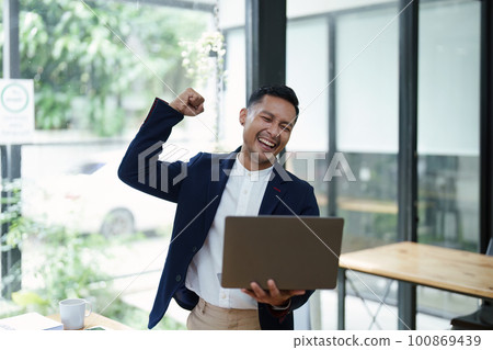 Portrait of an Asian male business owner standing with a computer showing happiness after a successful investment. Portrait of an Asian male business owner standing with a computer showing happiness after a successful investment. 100869439