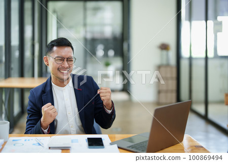 Portrait of an Asian male business owner standing with a computer showing happiness after a successful investment. Portrait of an Asian male business owner standing with a computer showing happiness after a successful investment. 100869494