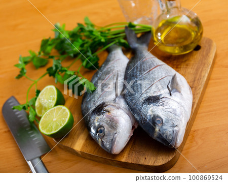 Two gilt-head breams laid on wooden cutting board with lime herbs salt and oil 100869524
