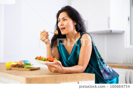 Woman leaning on kitchen table and eating salad Woman leaning on kitchen table and eating salad 100869754