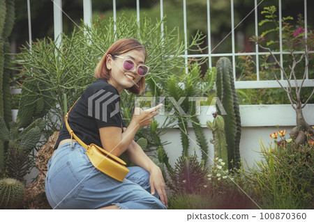 asian woman holding smartphone in hand toothy smiling face with happiness in beautiful home garden 100870360