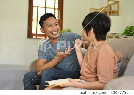 Joyful Asian dad telling funny story, laughing and playing with his son in the living room. 100872591